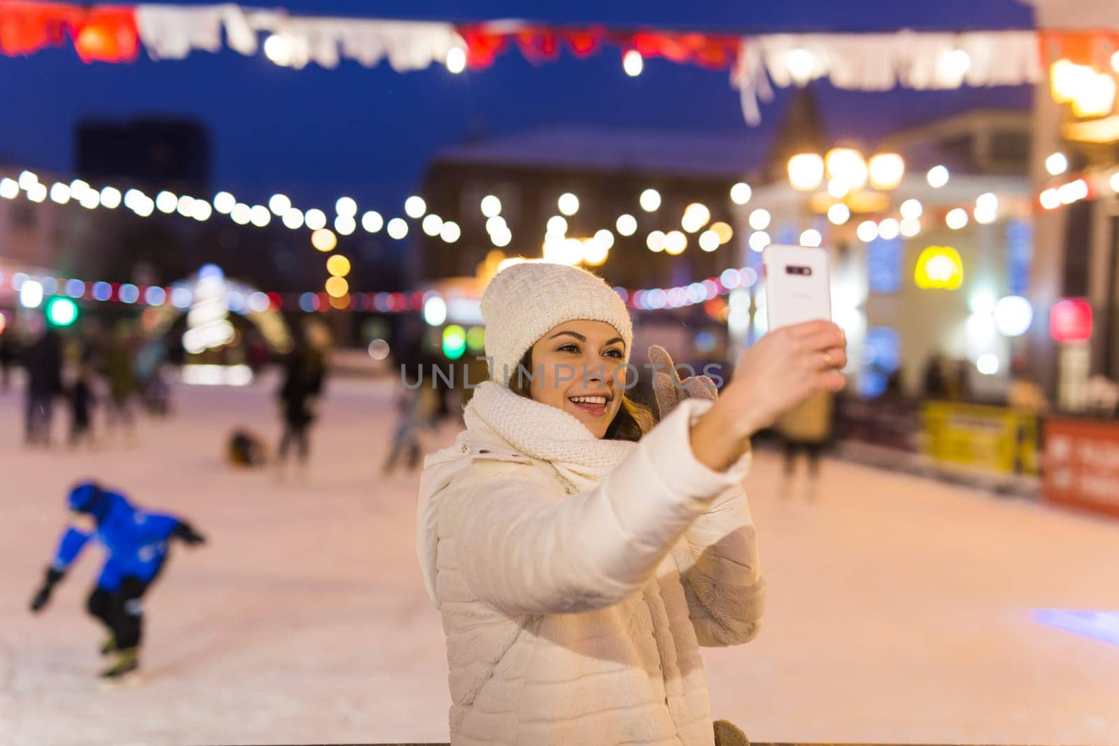 Christmas, winter, technology and leisure concept - happy young woman taking picture with smartphone on ice skating rink.