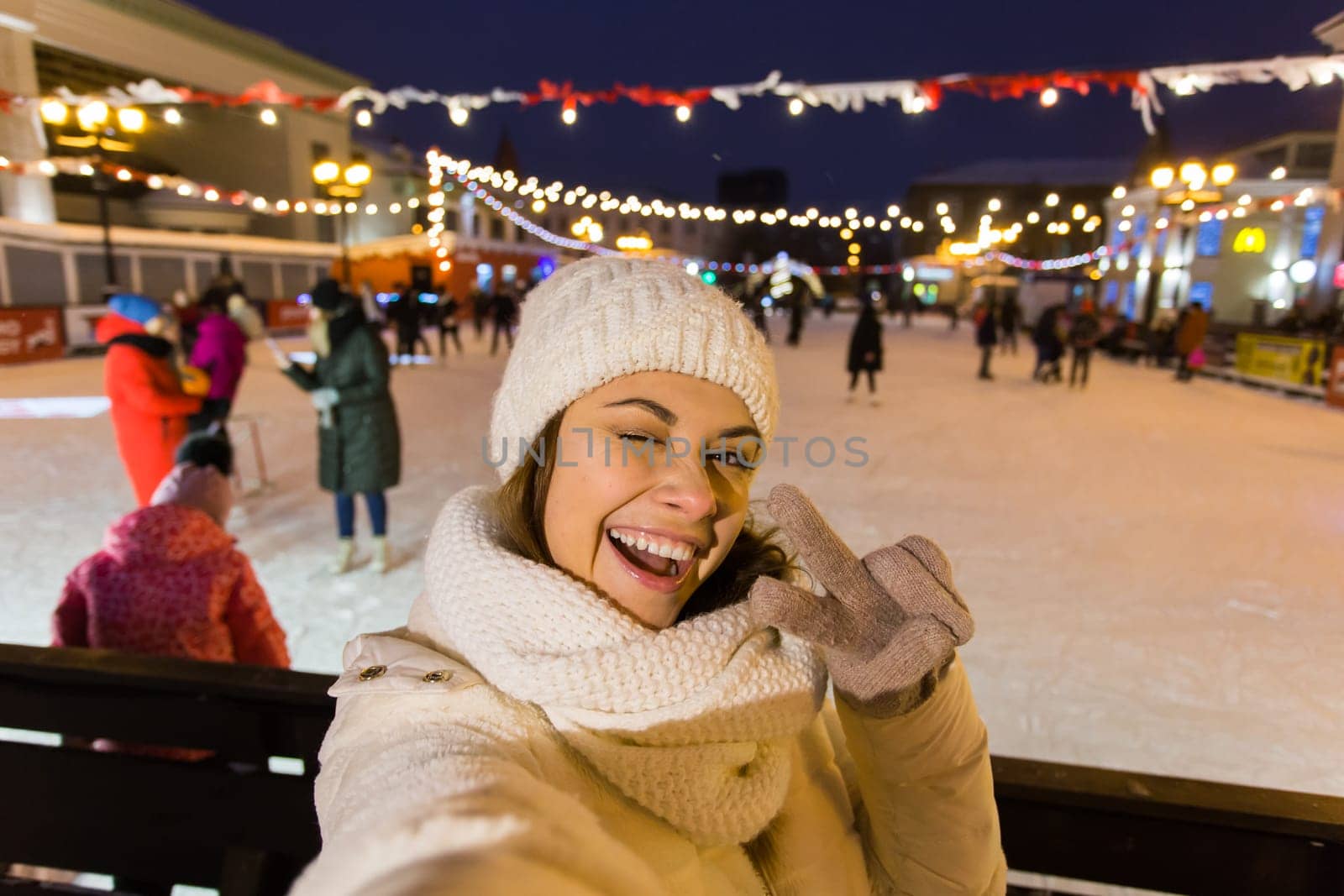 A woman on the rink is skating and taking selfie on smartphone. New Years Eve and Christmas. Fairy lights. Ice and snow mood concept. Winter