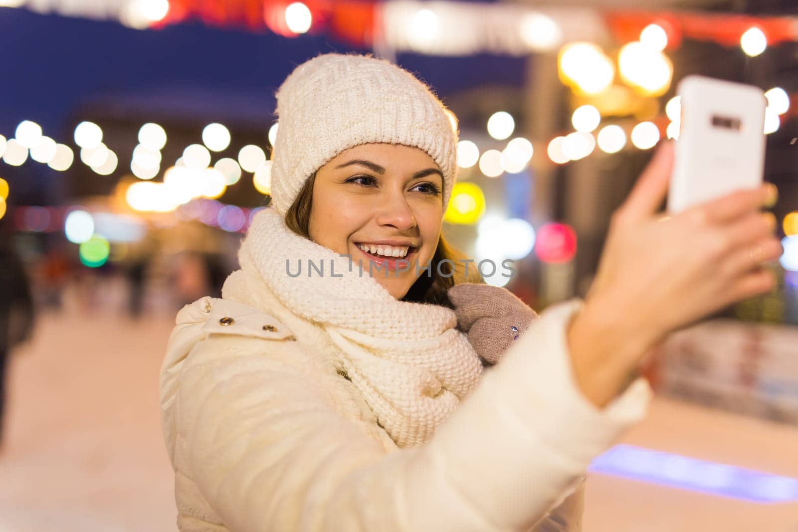 Christmas, winter, technology and leisure concept - happy young woman taking picture with smartphone on ice skating rink.