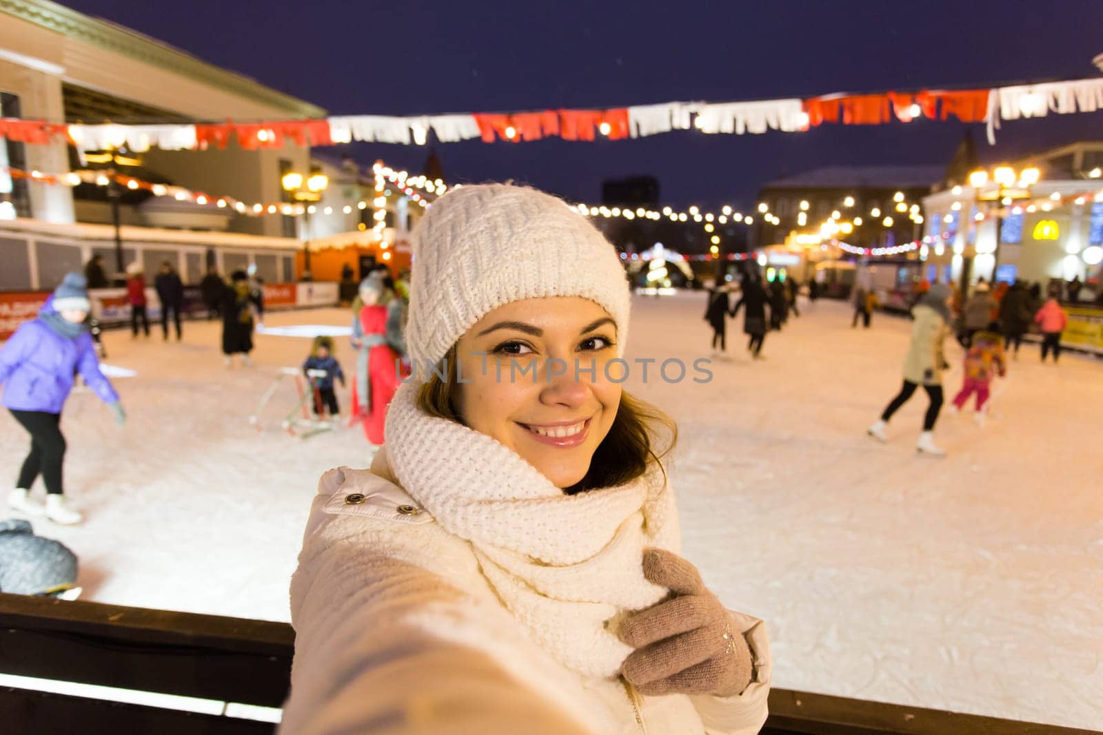 A woman on the rink is skating and taking selfie on smartphone. New Years Eve and Christmas. Fairy lights. Ice and snow mood concept. Winter