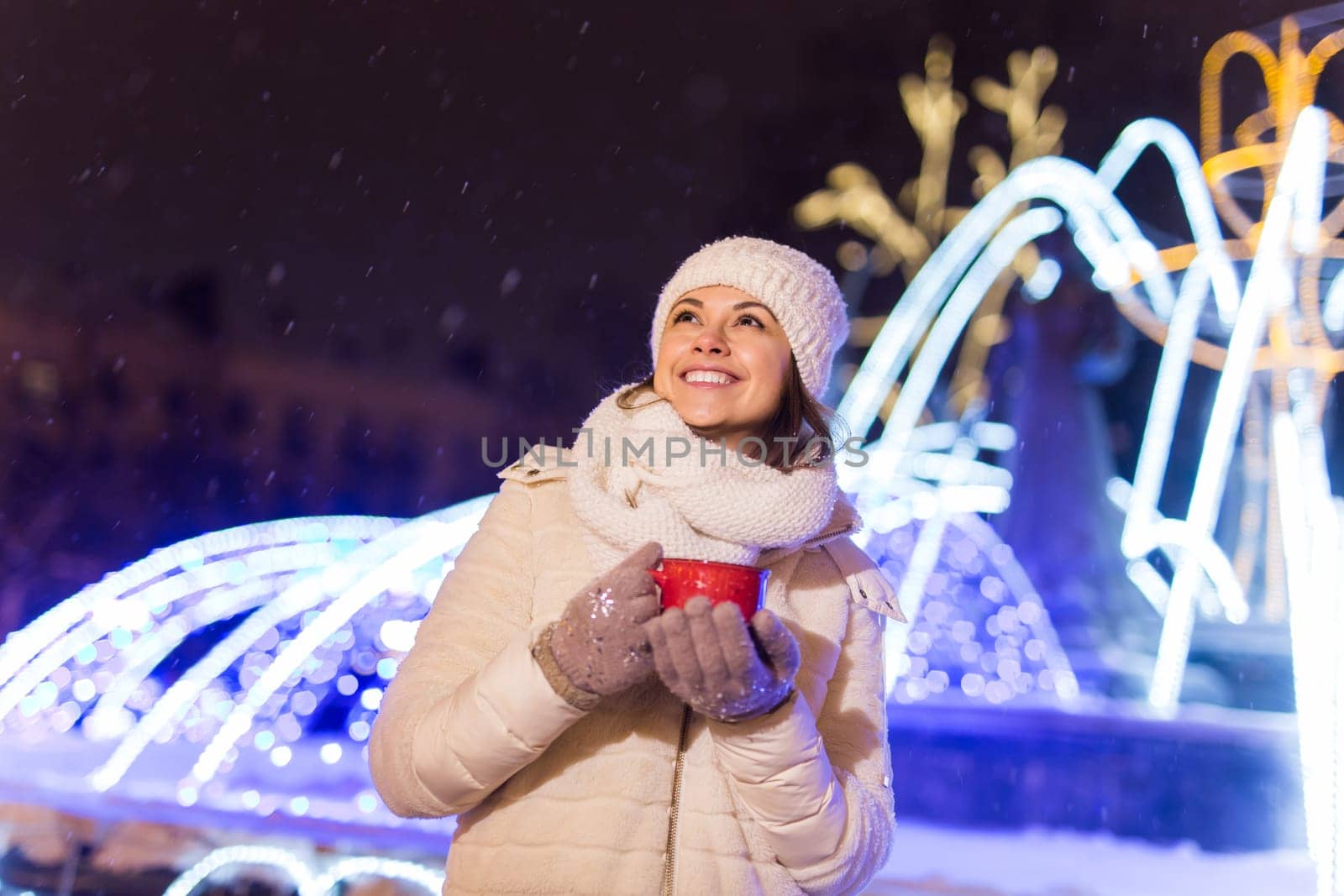 Girl standing over winter christmas city background snow snow drifts, stands warm jacket hat, holds cup tea with tea drinks.