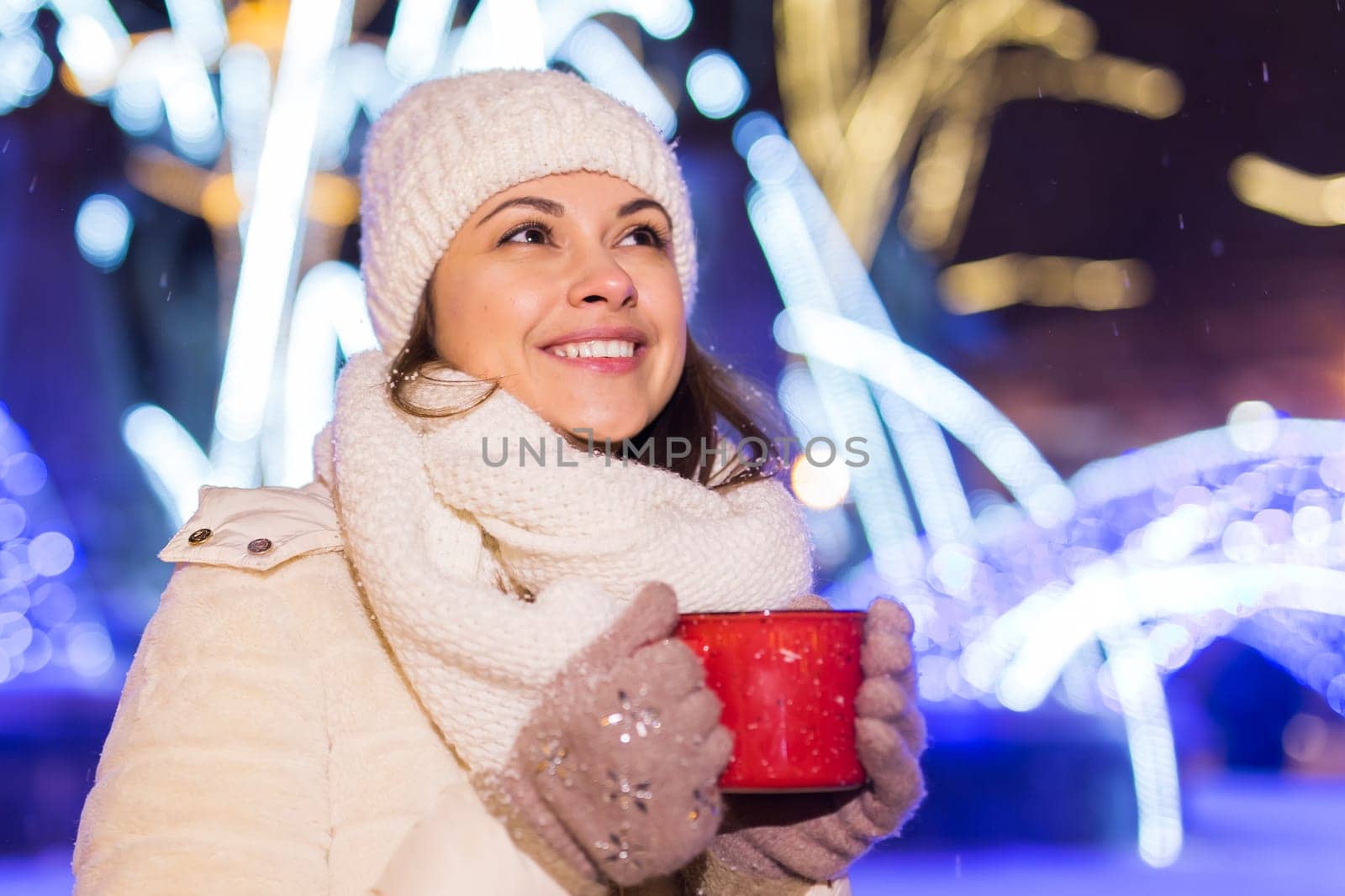 Girl standing over winter christmas city background snow snow drifts, stands warm jacket hat, holds cup tea with tea drinks.