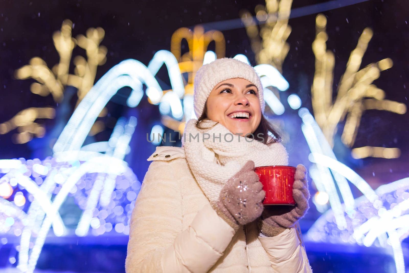 Girl standing over winter christmas city background snow snow drifts, stands warm jacket hat, holds cup tea with tea drinks.