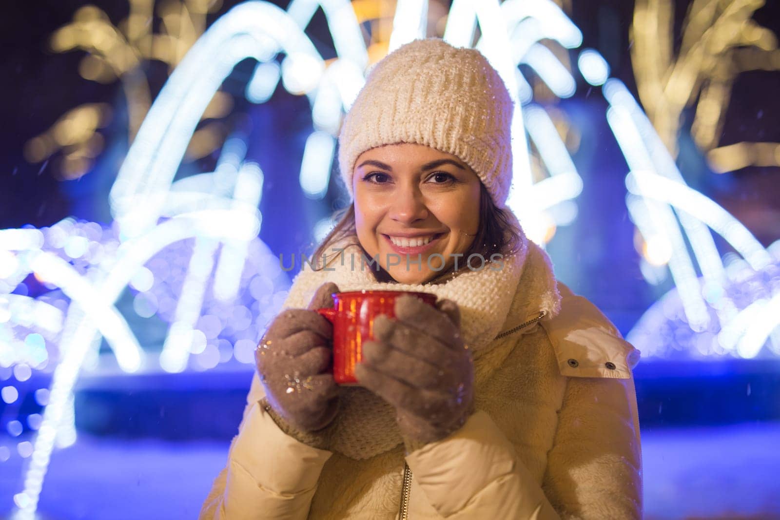 Girl standing over winter christmas city background snow snow drifts, stands warm jacket hat, holds cup tea with tea drinks.