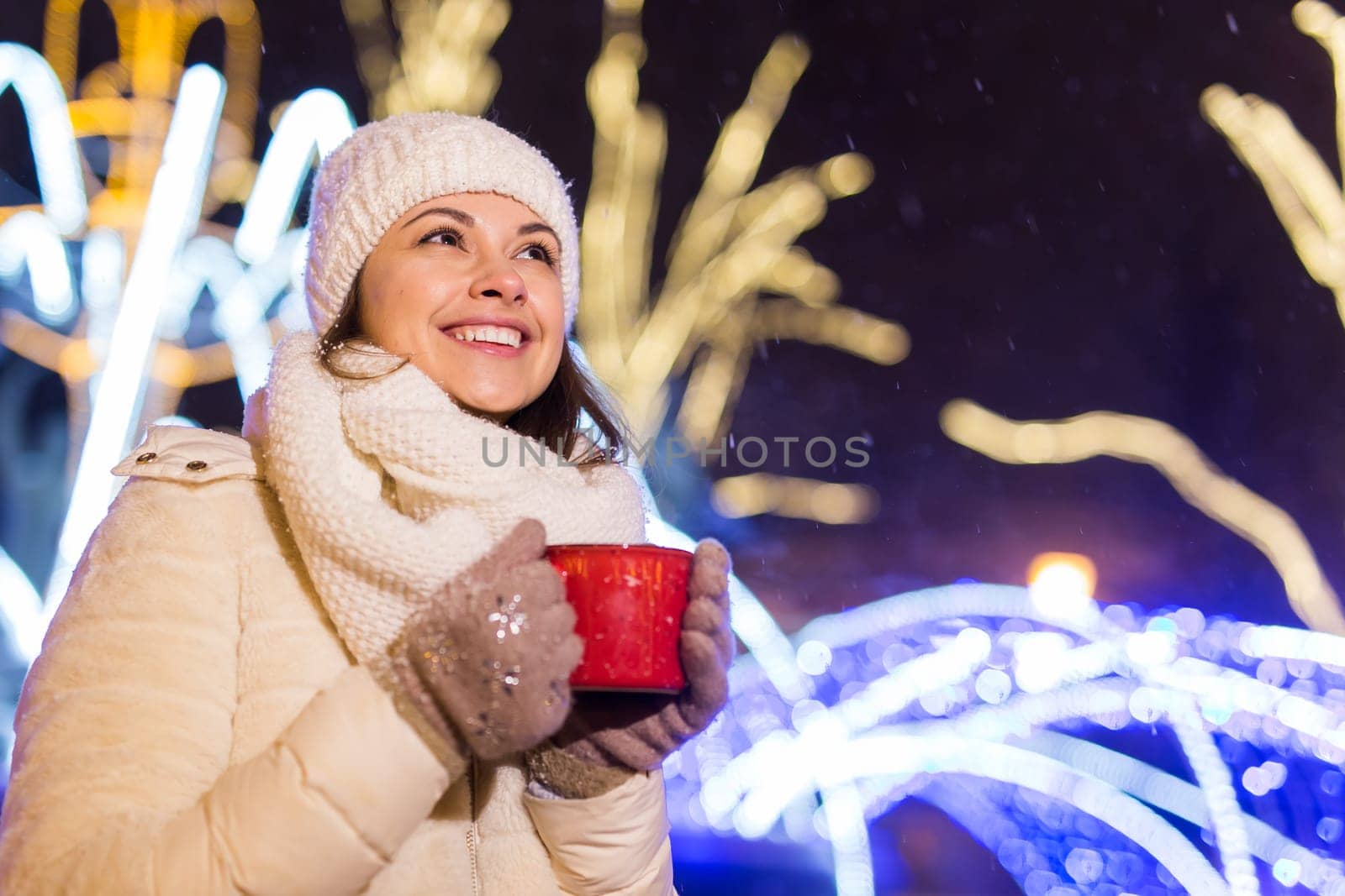 Girl standing over winter christmas city background snow snow drifts, stands warm jacket hat, holds cup tea with tea drinks.