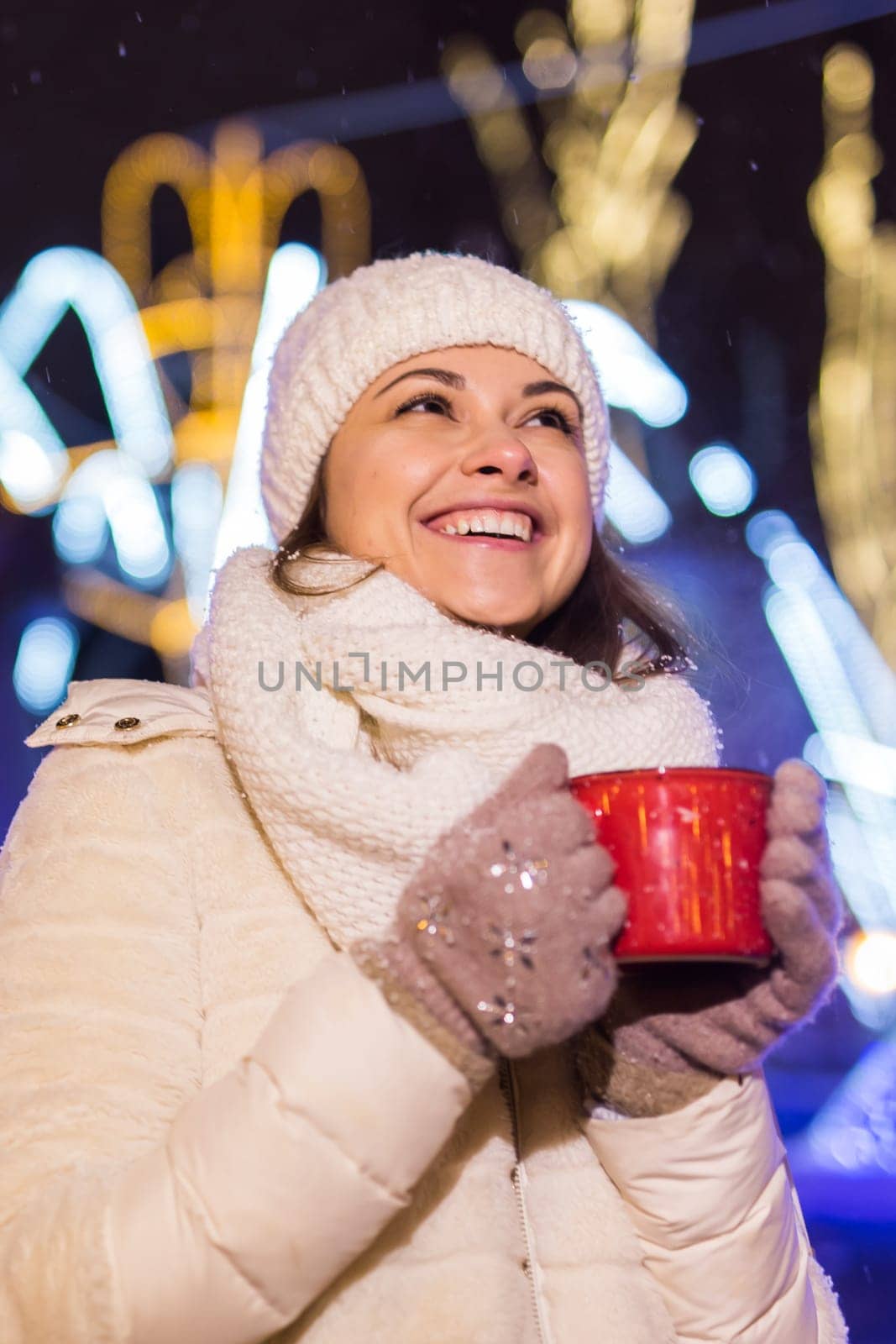 Girl standing over winter christmas city background snow snow drifts, stands warm jacket hat, holds cup tea with tea drinks.