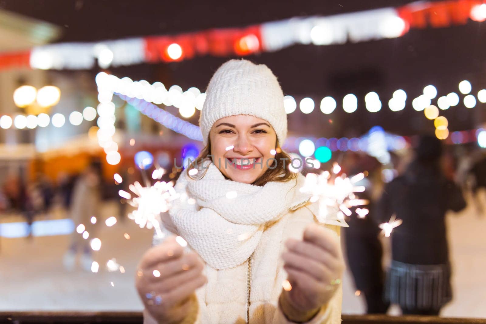 Young beautiful woman in knitted hat and scarf standing in the city with bengal light, sparkler. Concept celebration and christmas