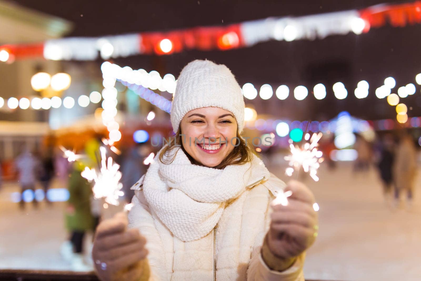 young woman wearing winter knitted clothes holding sparkler outdoors over snow background. Christmas holidays.