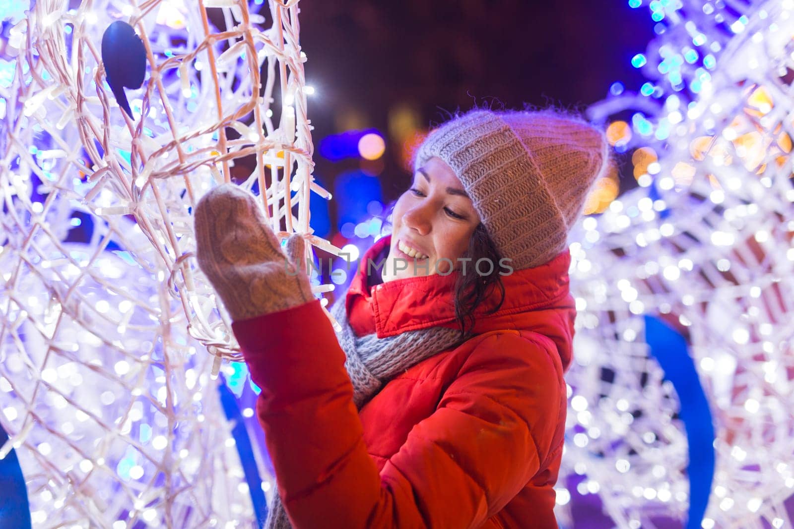 Girl having fun christmas decoration lights street. Young happy smiling woman wearing stylish knitted scarf and jacket outdoors. Model laughing. Winter wonderland city scene, New Year party.