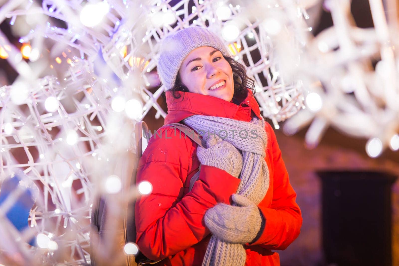 Girl having fun christmas decoration lights street. Young happy smiling woman wearing stylish knitted scarf and jacket outdoors. Model laughing. Winter wonderland city scene, New Year party.