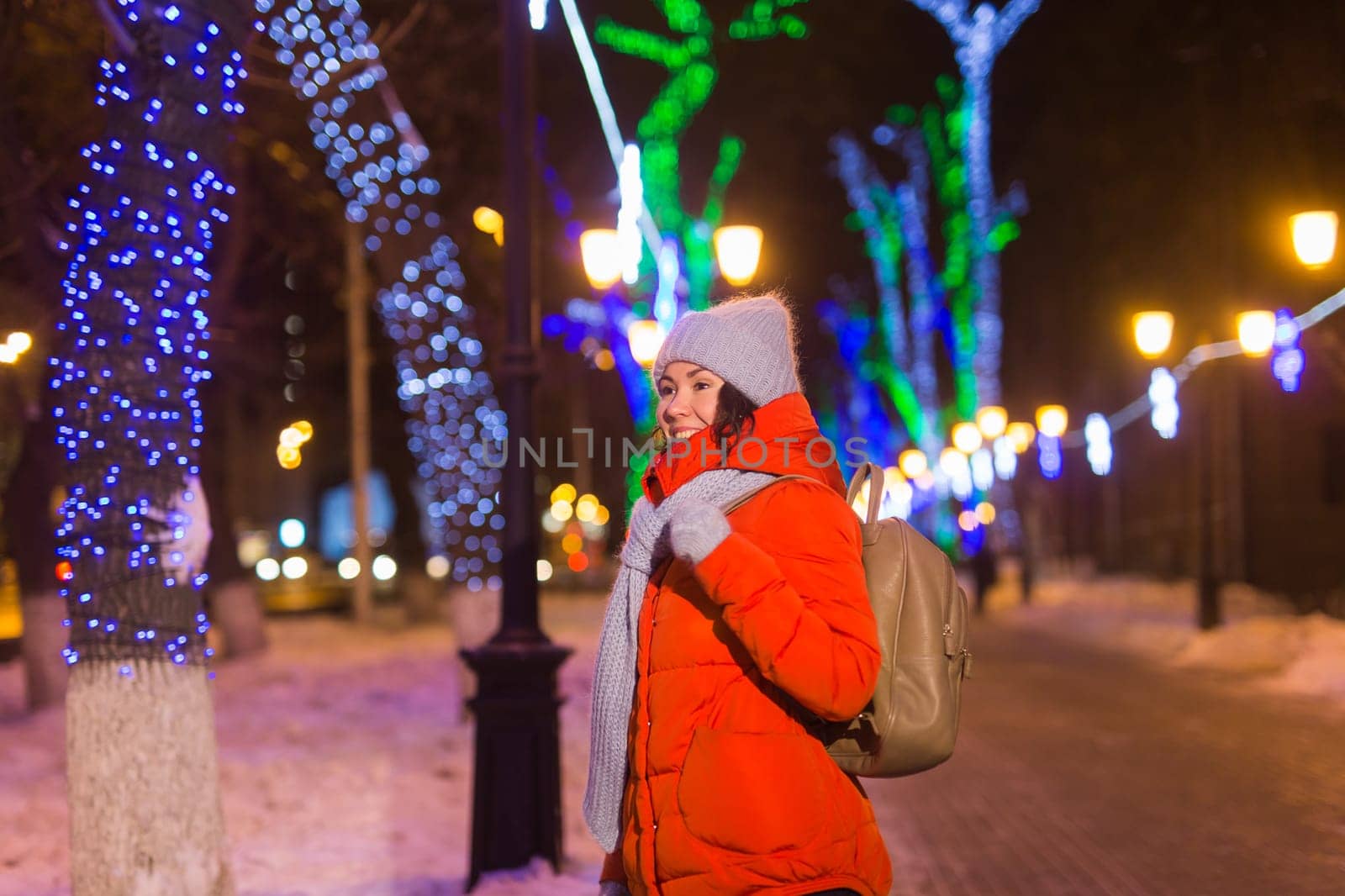 Girl having fun christmas decoration lights street. Young happy smiling woman wearing stylish knitted scarf and jacket outdoors. Model laughing. Winter wonderland city scene, New Year party.