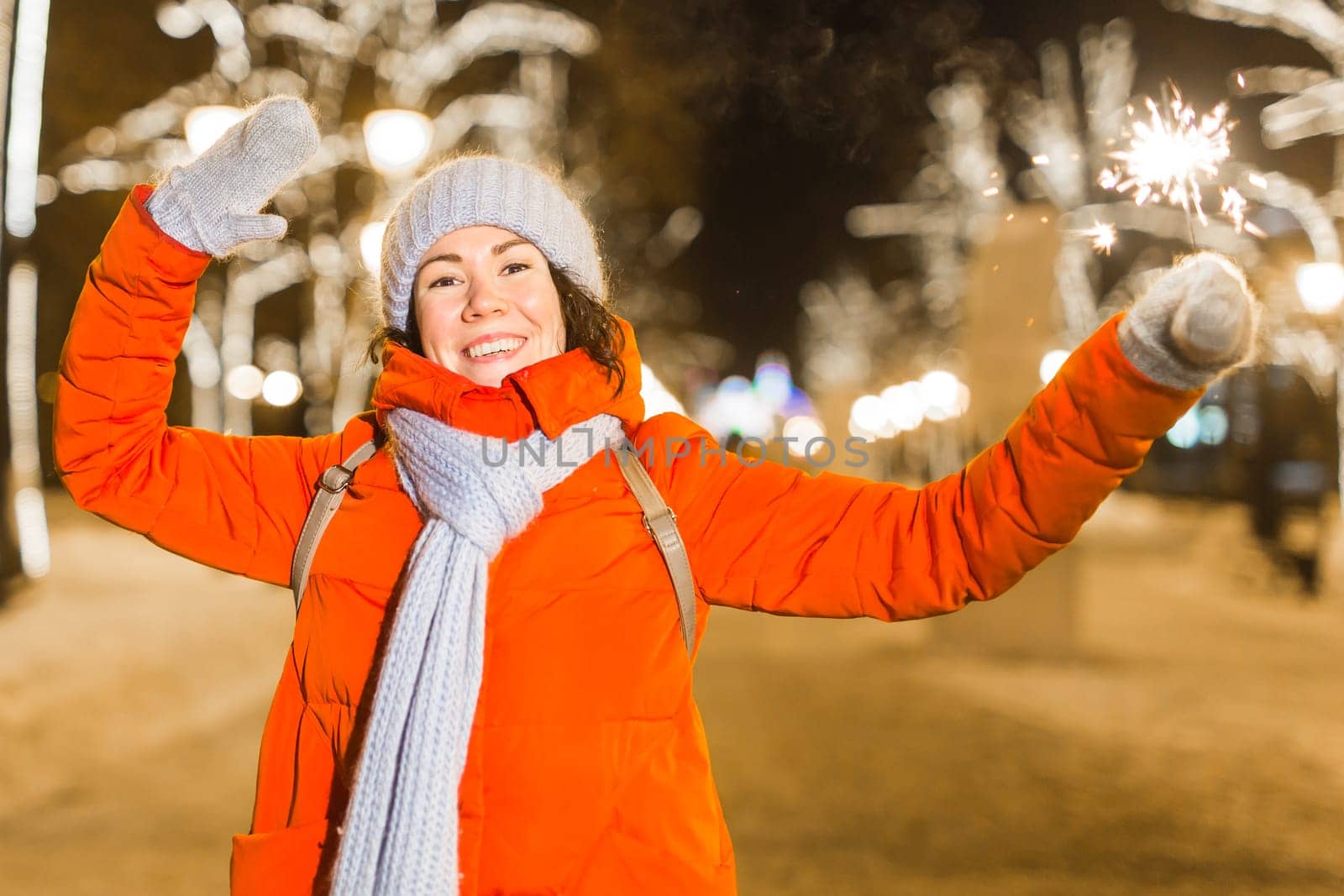 Young beautiful woman in knitted hat and scarf standing in the city with bengal light, sparkler. Concept celebration and christmas
