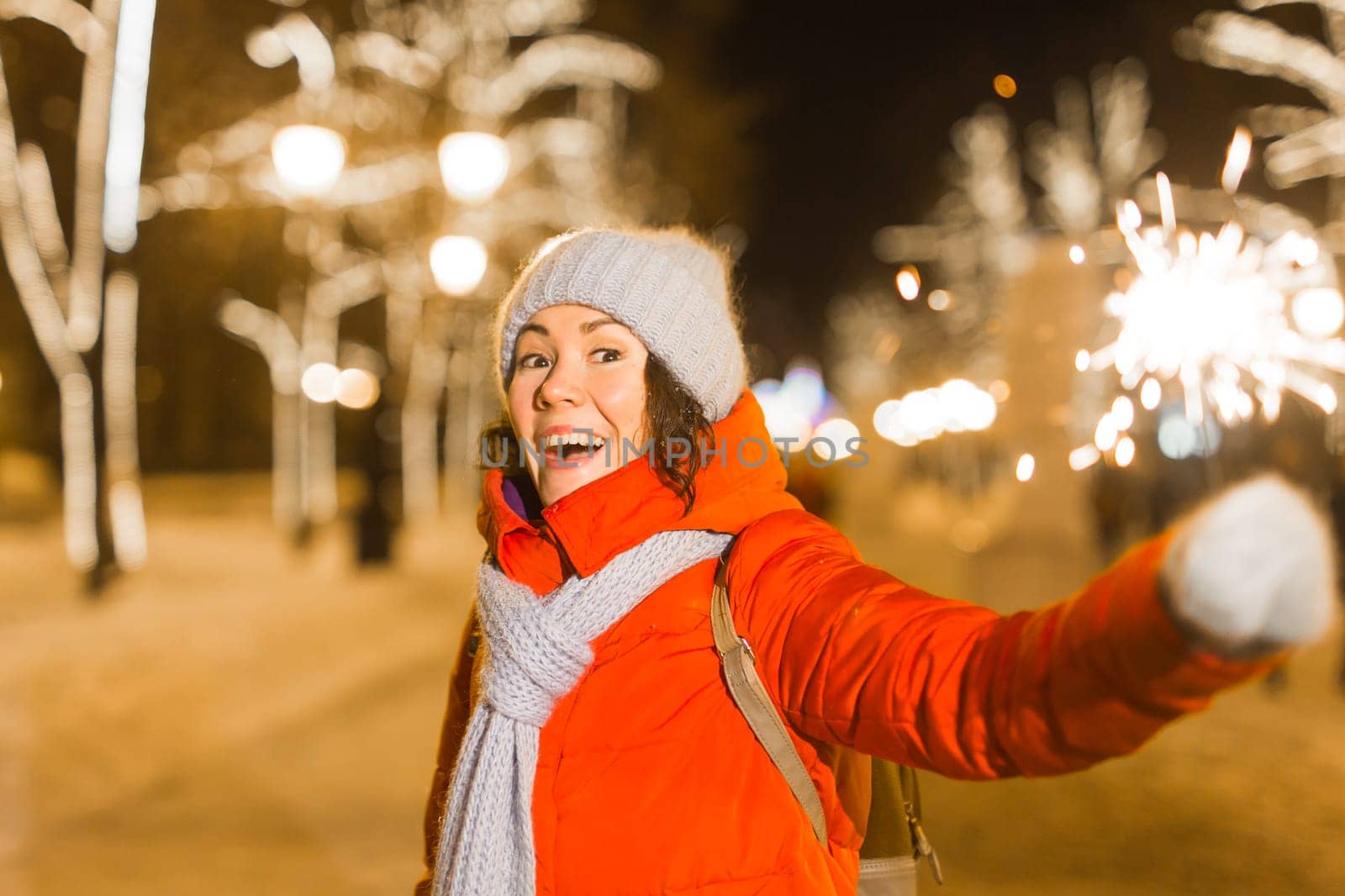 Young beautiful woman in knitted hat and scarf standing in the city with bengal light, sparkler. Concept celebration and christmas