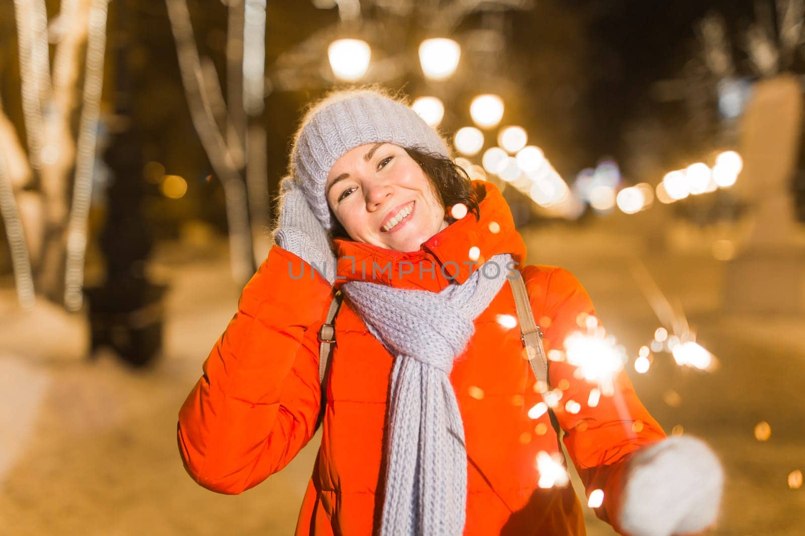 Young beautiful woman in knitted hat and scarf standing in the city with bengal light, sparkler. Concept celebration and christmas