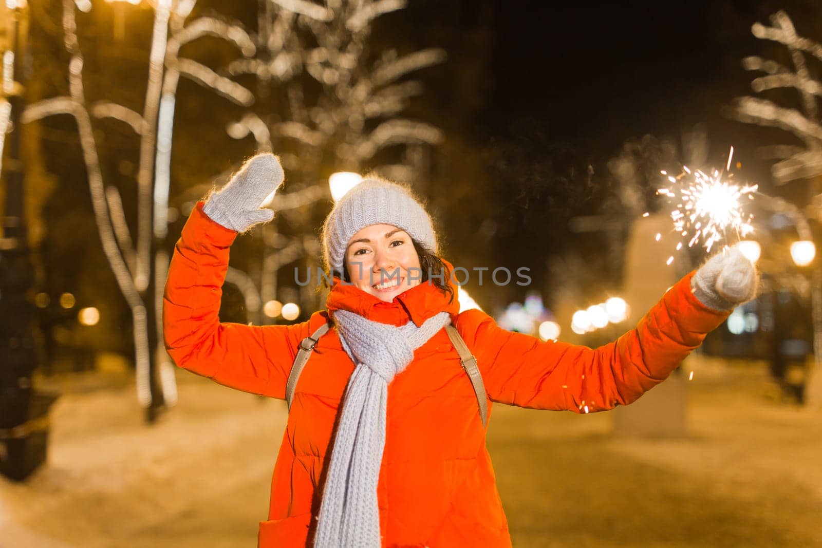 Young beautiful woman in knitted hat and scarf standing in the city with bengal light, sparkler. Concept celebration and christmas