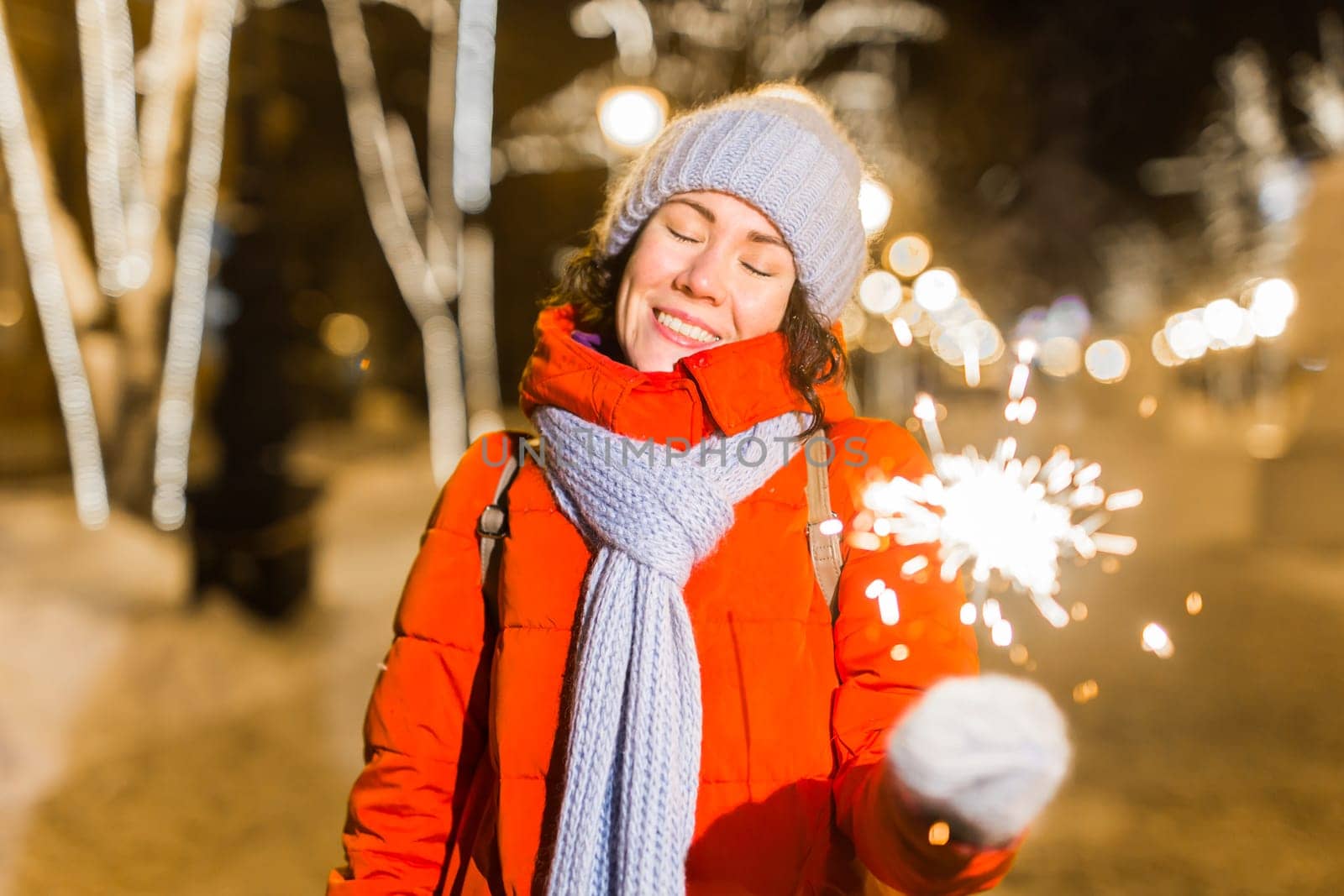 Young beautiful woman in knitted hat and scarf standing in the city with bengal light, sparkler. Concept celebration and christmas