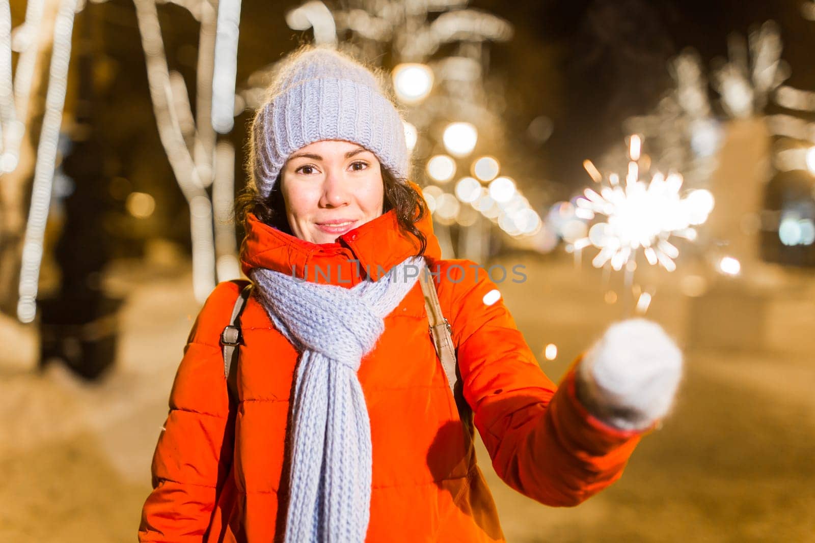 young woman wearing winter knitted clothes holding sparkler outdoors over snow background. Christmas holidays.