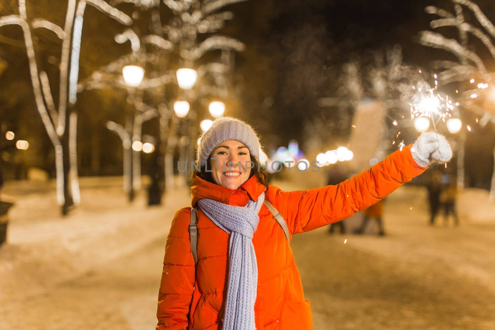 young woman wearing winter knitted clothes holding sparkler outdoors over snow background. Christmas holidays.