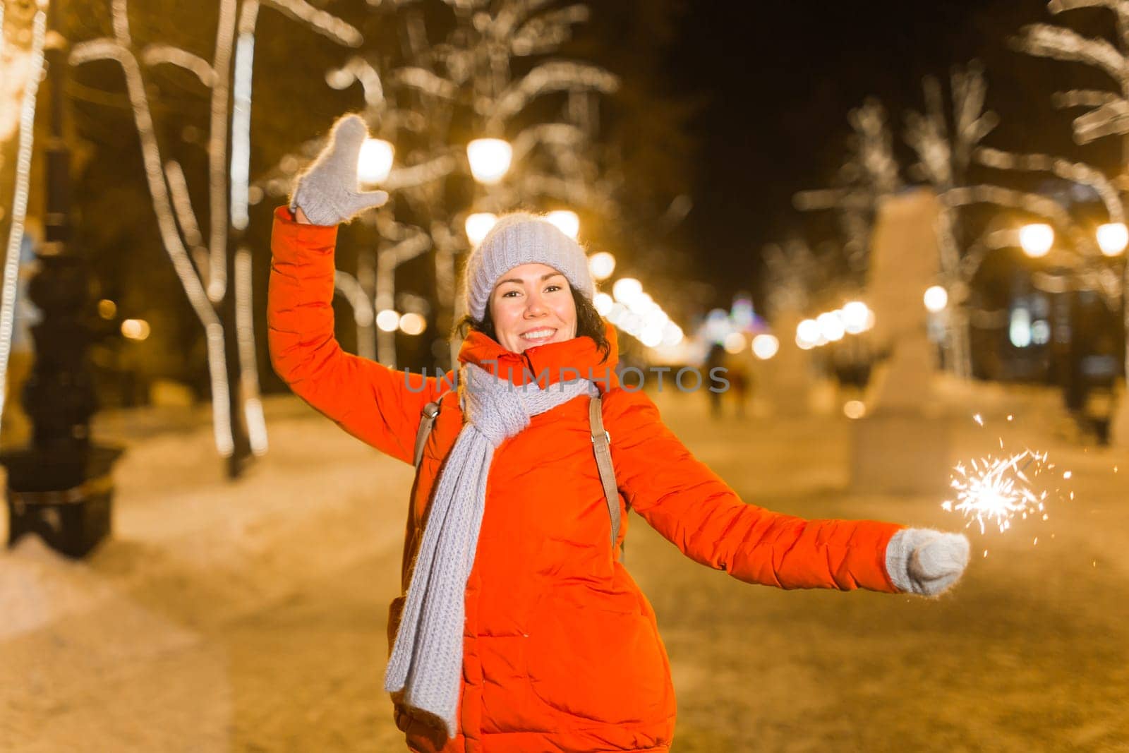 Young beautiful woman in knitted hat and scarf standing in the city with bengal light, sparkler. Concept celebration and christmas