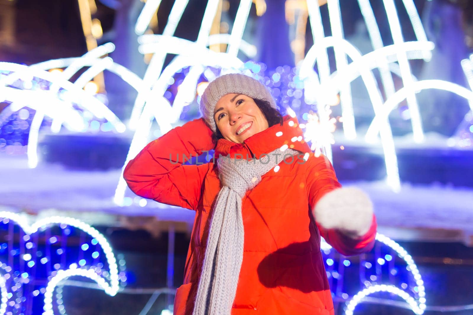 young woman wearing winter knitted clothes holding sparkler outdoors over snow background. Christmas holidays.