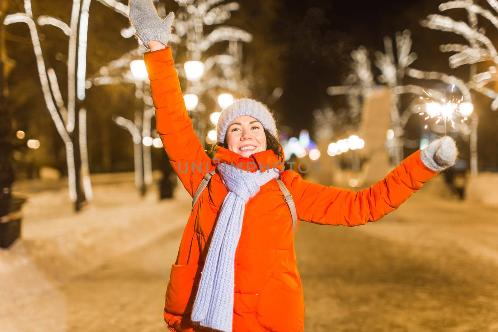 young woman wearing winter knitted clothes holding sparkler outdoors over snow background. Christmas holidays.
