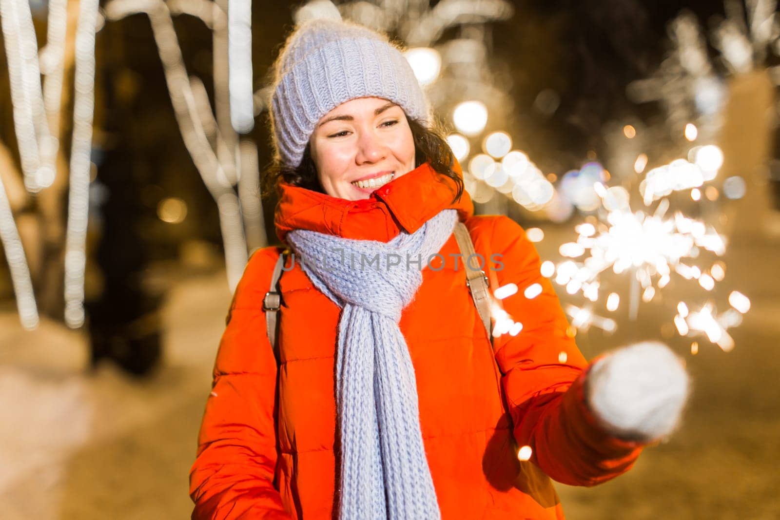 Young beautiful woman in knitted hat and scarf standing in the city with bengal light, sparkler. Concept celebration and christmas