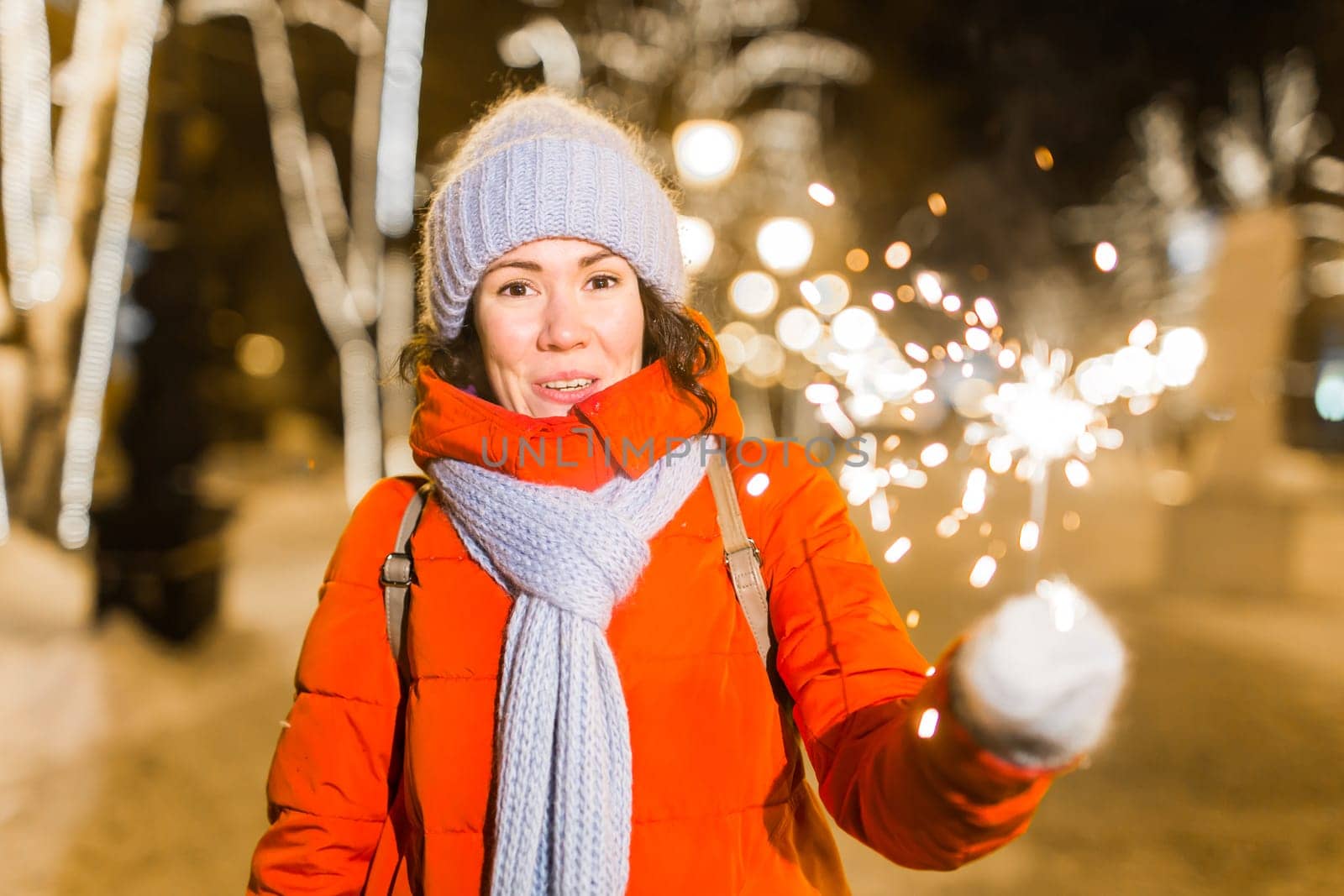 Young beautiful woman in knitted hat and scarf standing in the city with bengal light, sparkler. Concept celebration and christmas