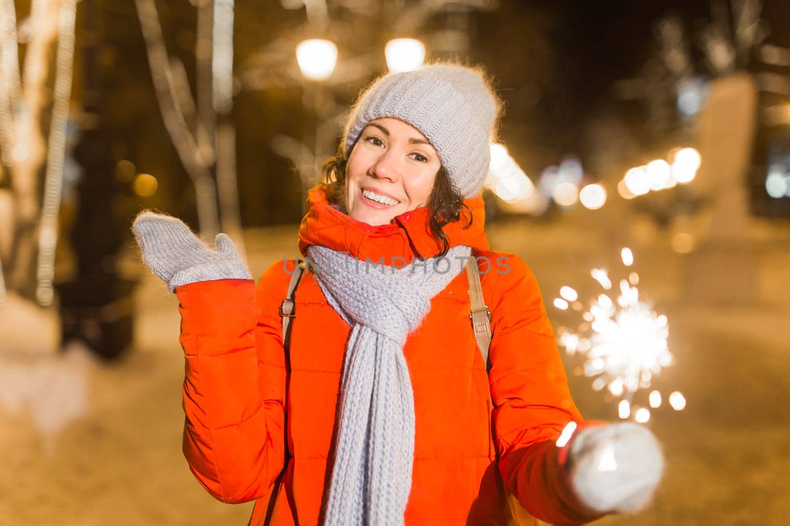 young woman wearing winter knitted clothes holding sparkler outdoors over snow background. Christmas holidays.