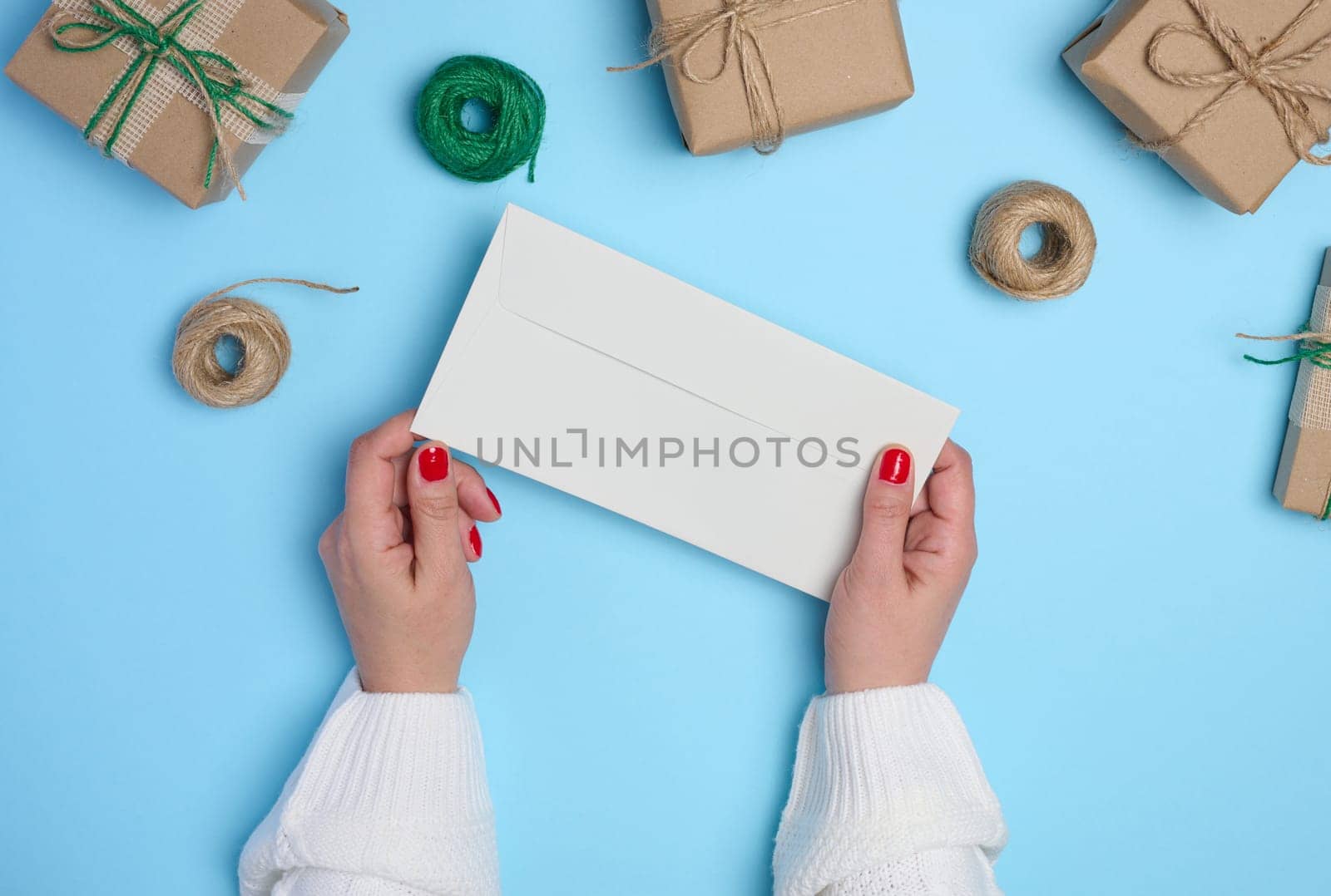 Female hands holding a white paper envelope on a blue background, around a box with gifts, top view