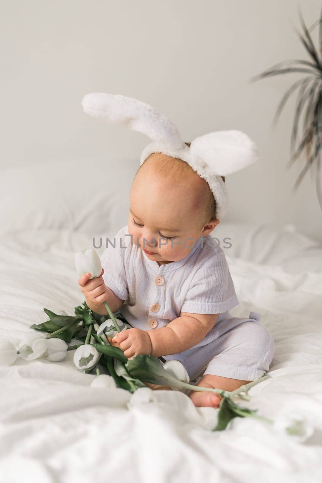 Cute funny baby with bunny ears, with a tulips at home on a bed of white bedding. Easter greeting card.