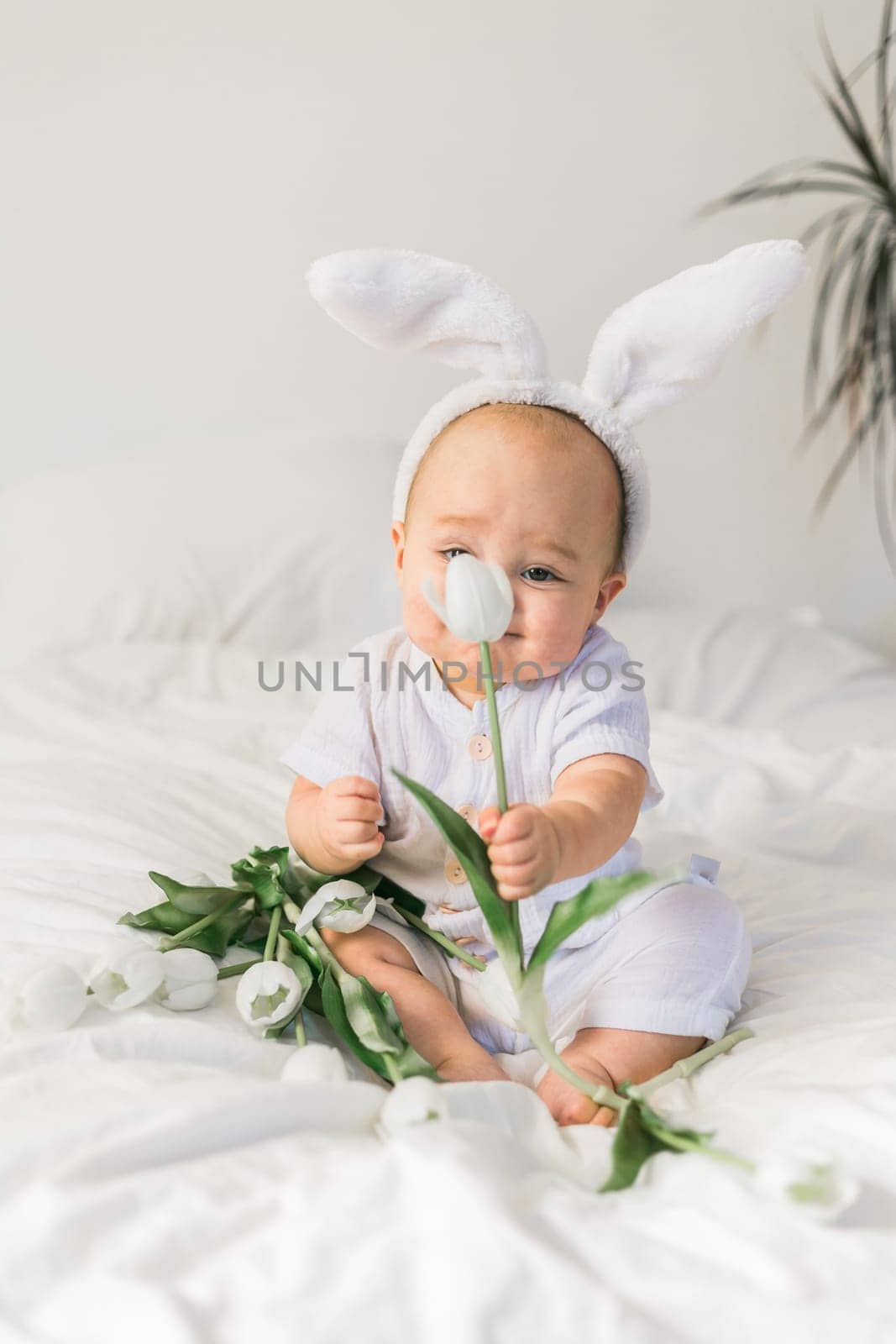 Cute funny baby with bunny ears, with a tulips at home on a bed of white bedding. Easter greeting card.