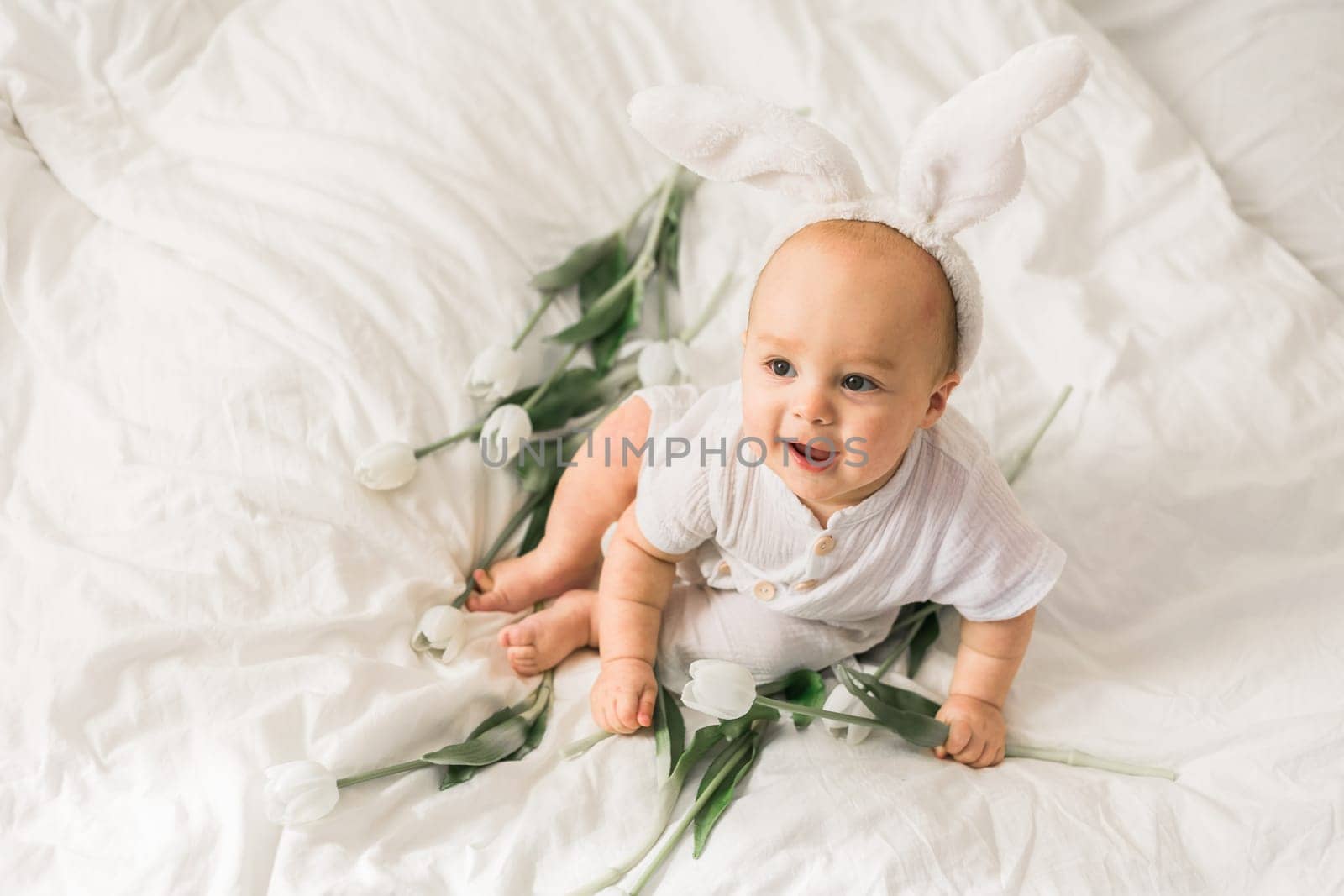 Cute funny baby with bunny ears, with a tulips at home on a bed of white bedding. Easter greeting card.