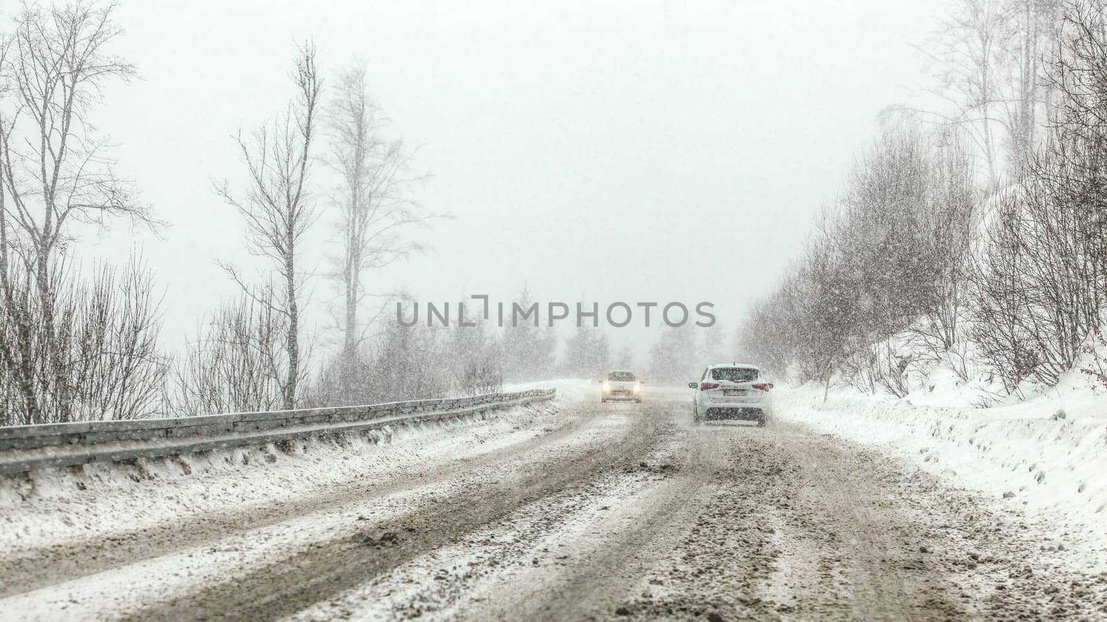Cars driving on snow covered forest road during snowstorm, fog in disance, view from car behind. Dangerous driving conditions.