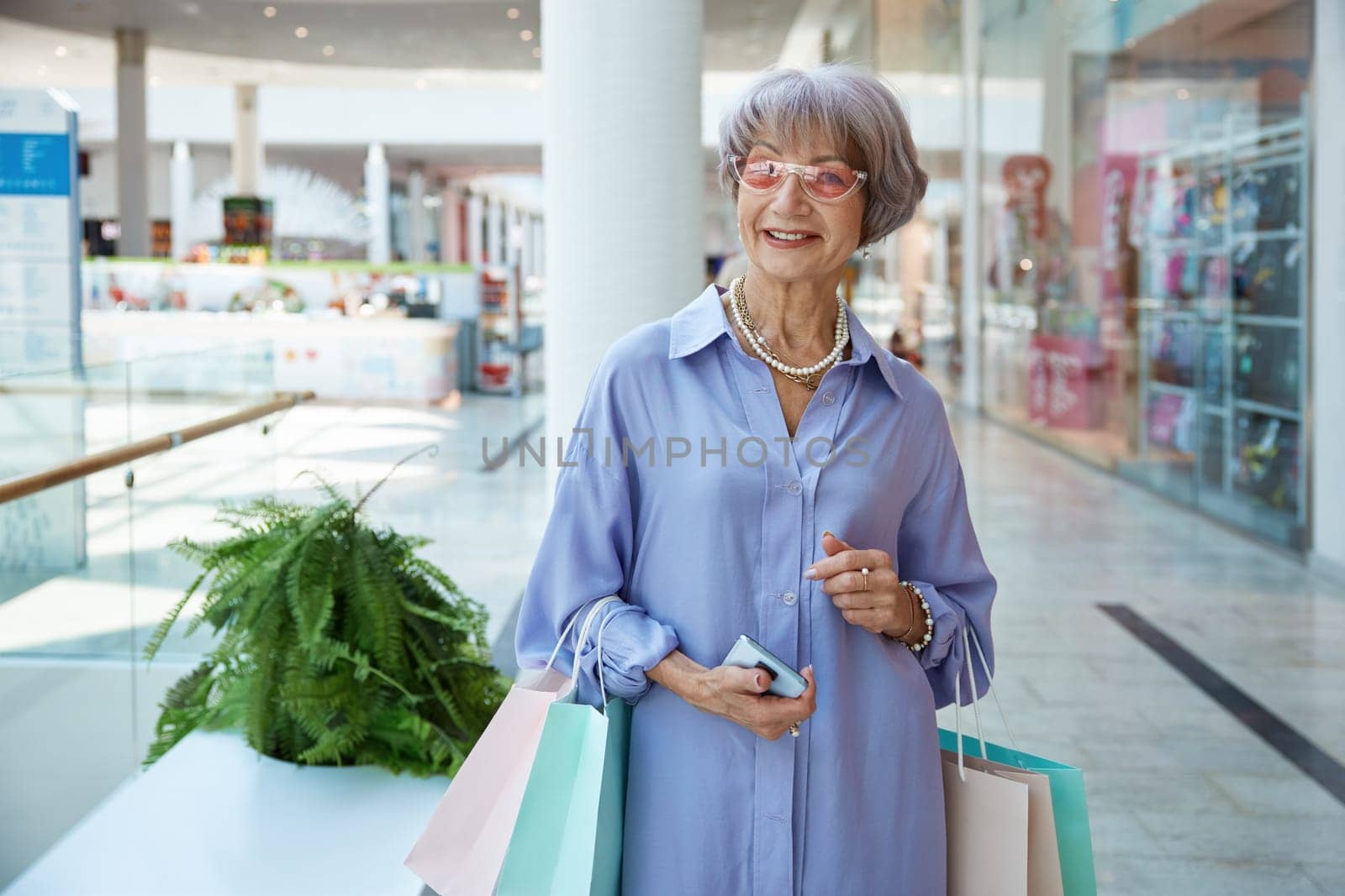 Happy attractive senior woman with shopping bags over mall store showcase. Stylish grandmother feeling satisfied enjoying discounts in shop