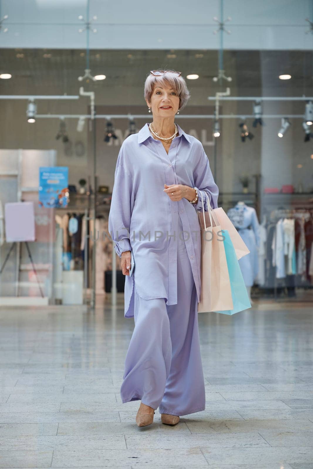 Full length portrait of attractive senior woman with shopping bags over store showcase. Stylish grandmother feeling satisfied enjoying discounts in shop
