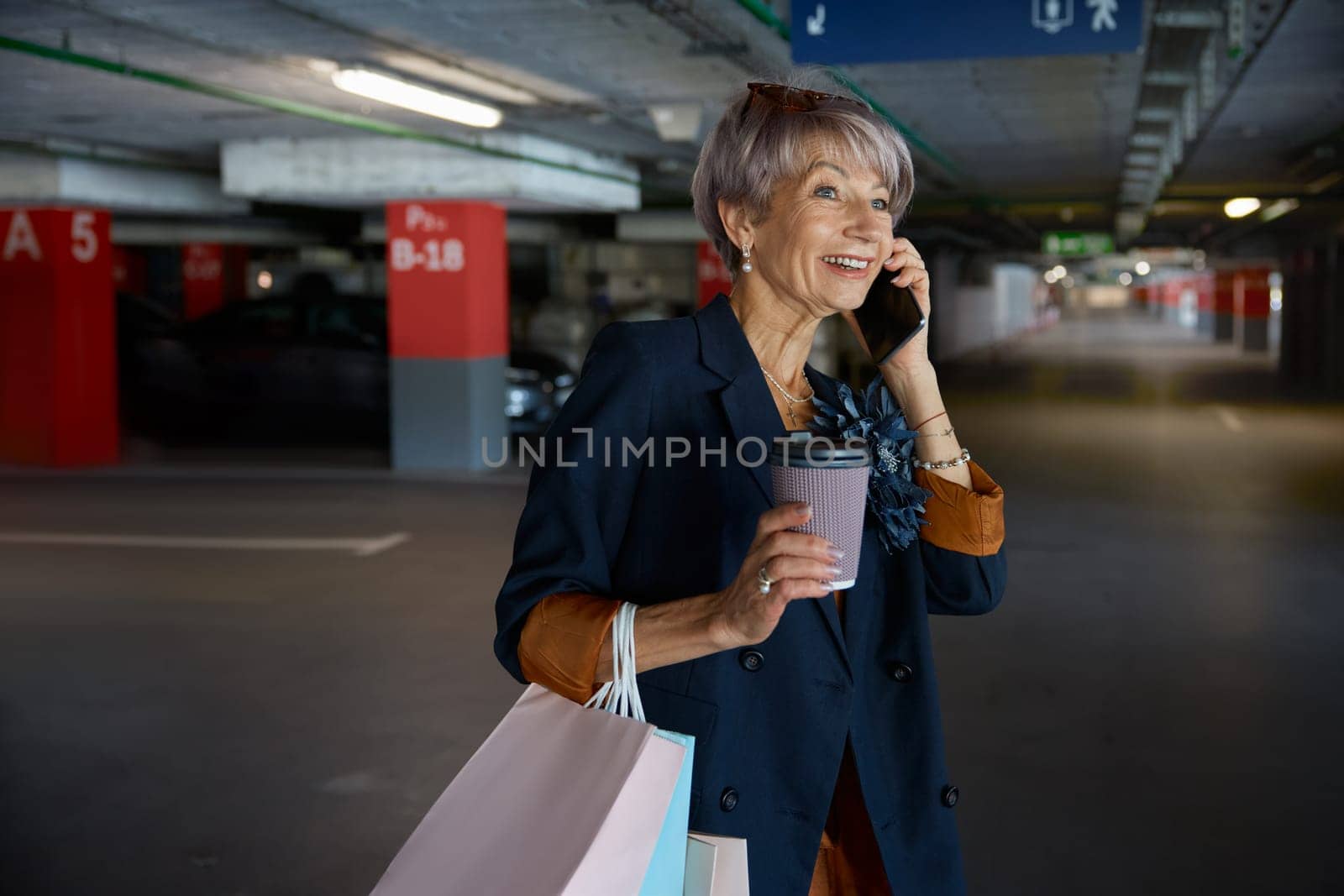 Happy elderly woman talking mobile phone walking through parking of shopping mall. Senior lady sharing expression after successful purchases with low prices