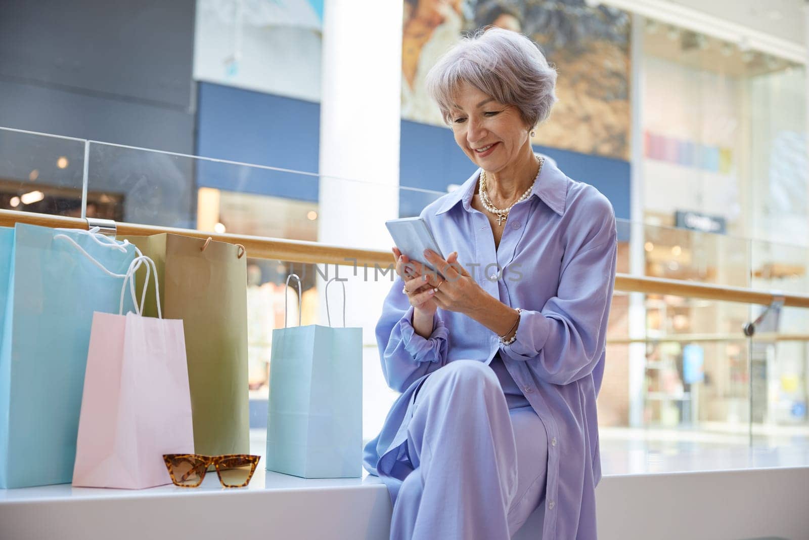 Beautiful mature woman resting after shopping sitting on bench in supermarket and chatting social media with friends or calling taxi cab using smartphone