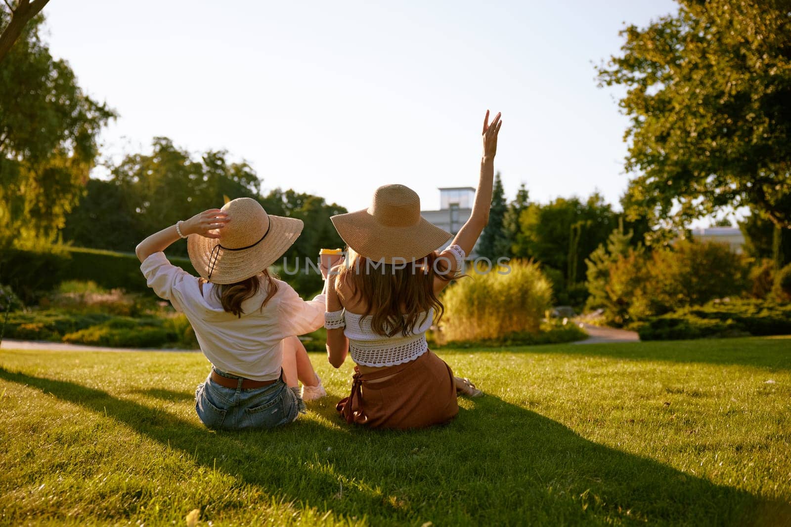 Back view of two pretty girls best friends in park. Teenage female sitting on grass meadow in ciry park