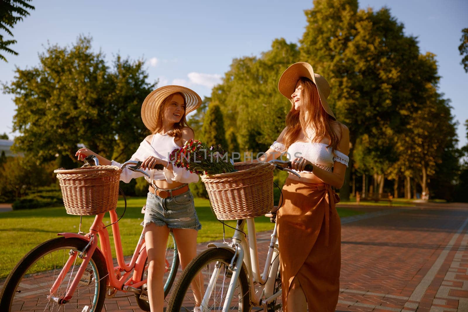 Two beautiful girl best friends having nice conversation while walking with bicycle at public park. Charming young female florist enjoying company of each other