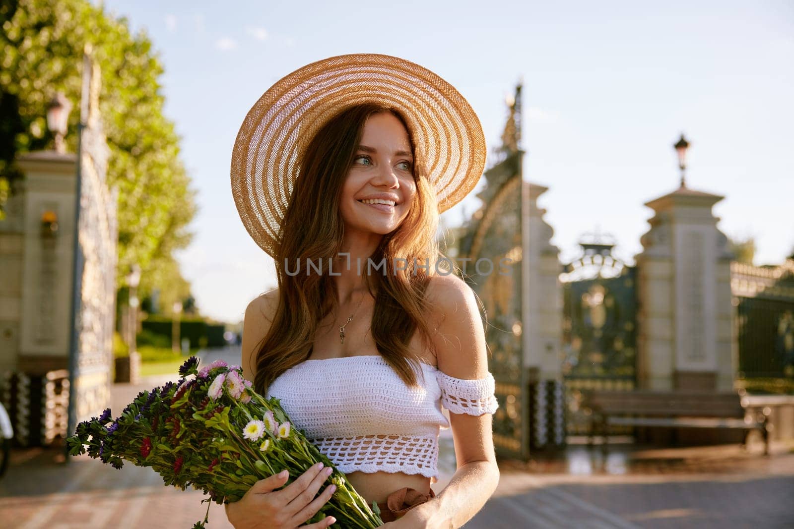 Portrait of gentle beautiful young woman holding wildflower bouquet in summer park. Charming romantic girl in fashion straw hat