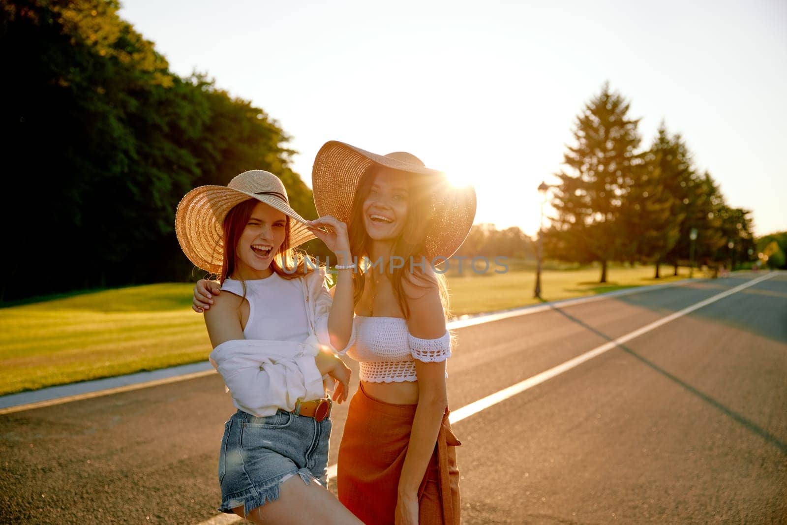 Happy young female friends posing for camera standing on road in sunset light beams. Romantic hipster girls with natural beauty enjoying travel and adventure feeling carefree