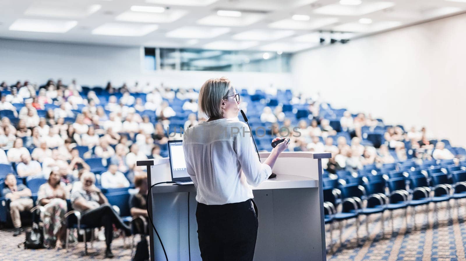 Female speaker giving a talk on corporate business conference. Unrecognizable people in audience at conference hall. Business and Entrepreneurship event