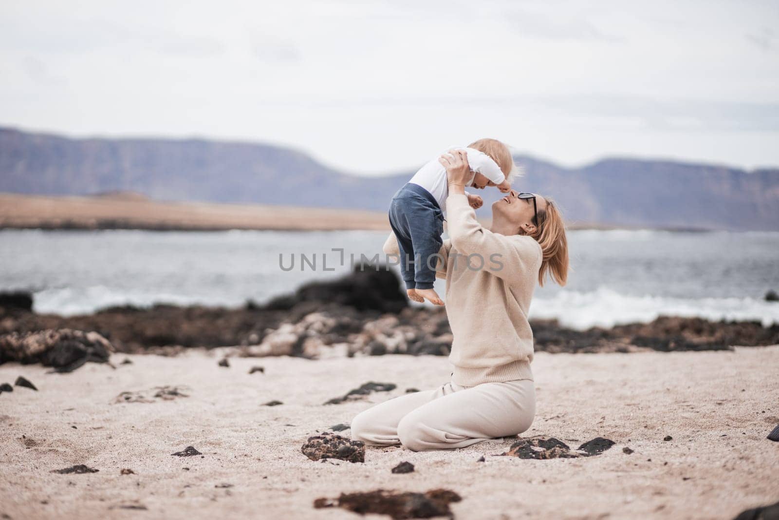 Mother enjoying winter vacations holding, playing and lifting his infant baby boy son high in the air on sandy beach on Lanzarote island, Spain. Family travel and vacations concept.