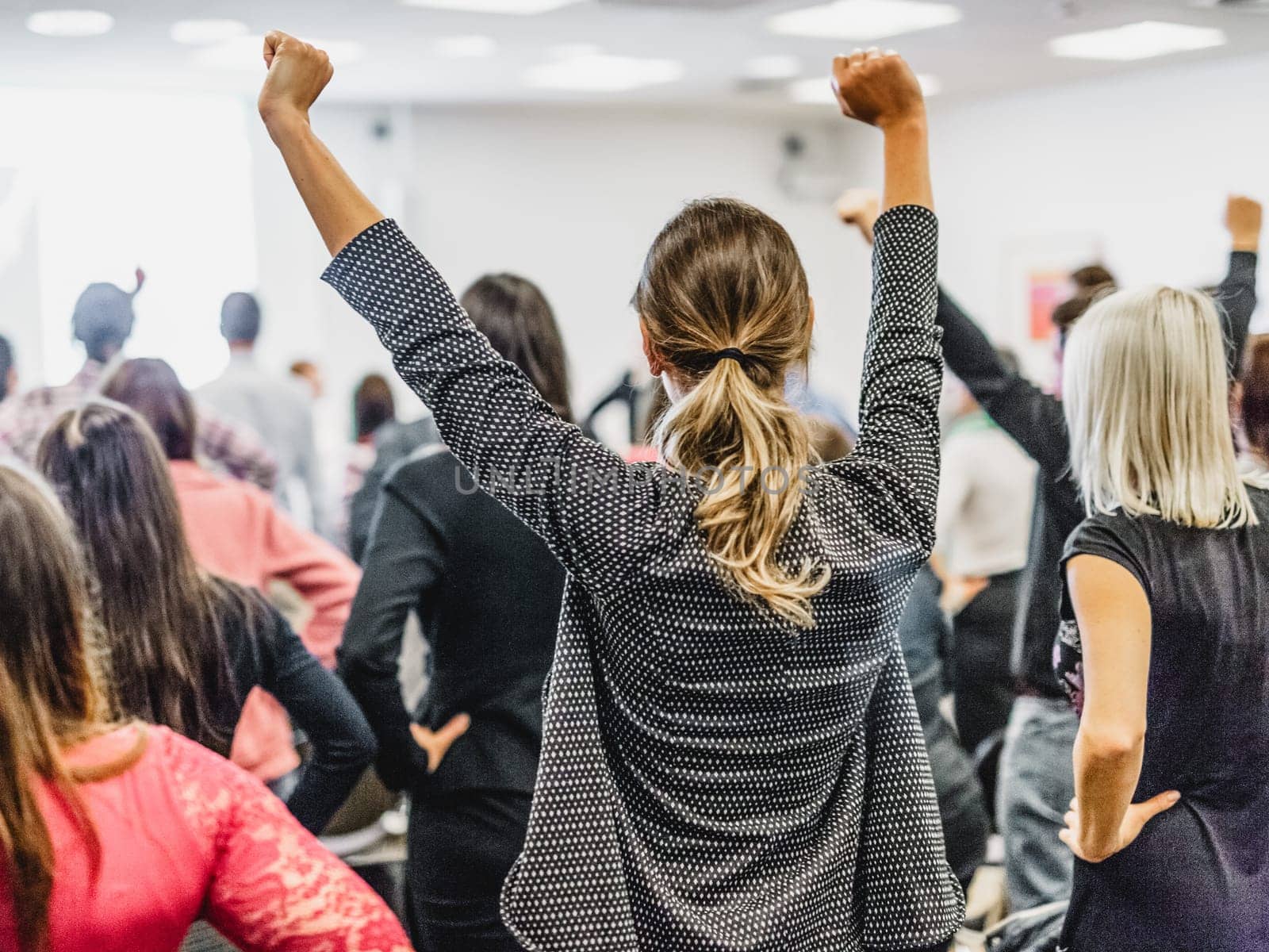 Life coaching symposium. Speaker giving interactive motivational speech at business workshop. Rear view of unrecognizable participants feeling empowered and motivated, hands raised high in air.