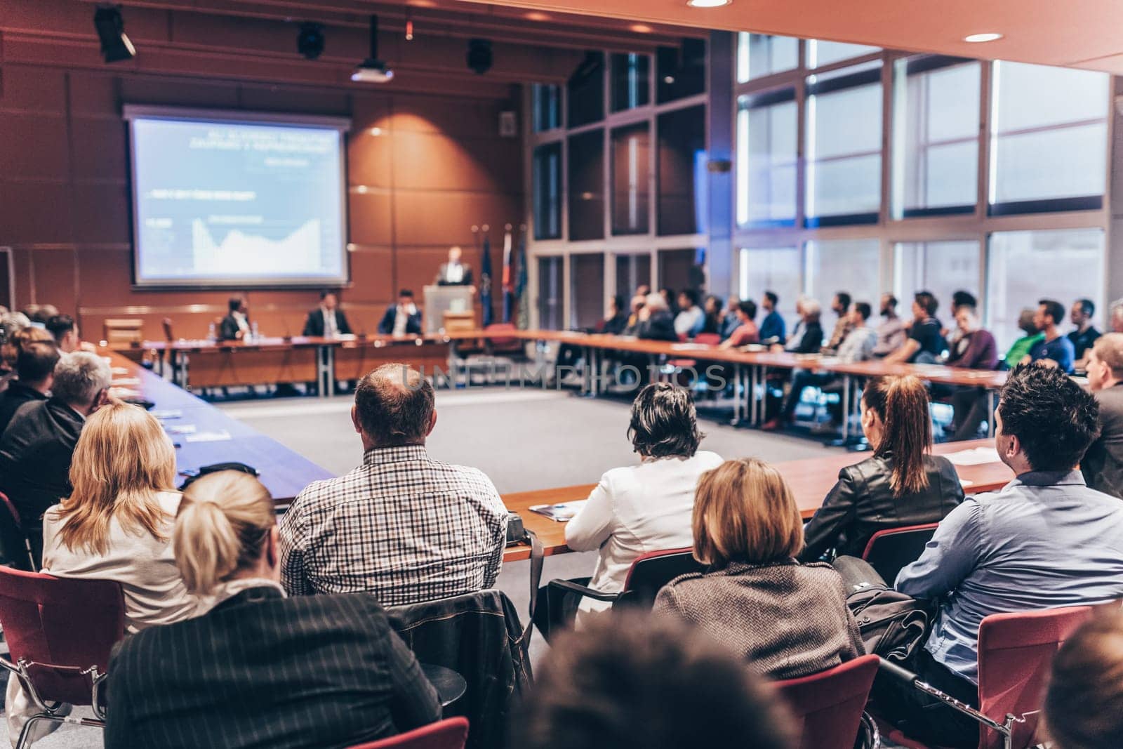 Speaker giving a talk on corporate business event. Audience at the conference hall. Business and Entrepreneurship event. Focus on unrecognizable business people in the rear.