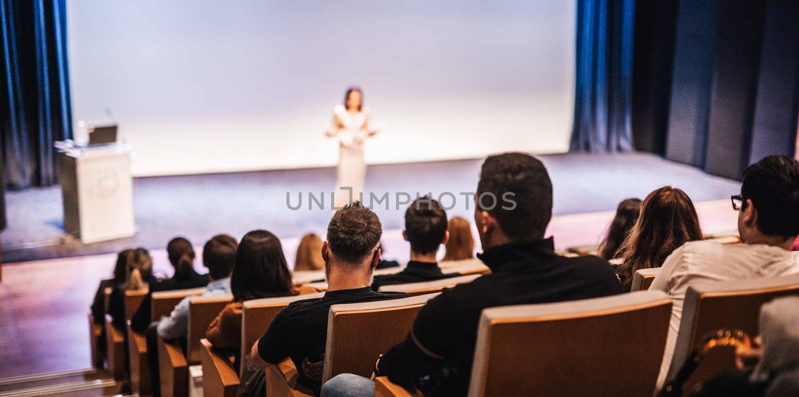 Business and entrepreneurship symposium. Female speaker giving a talk at business meeting. Audience in conference hall. Rear view of unrecognized participant in audience.