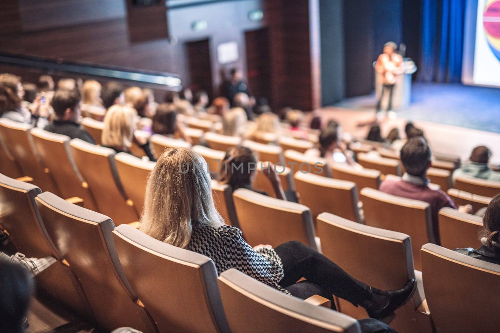 Business and entrepreneurship symposium. Female speaker giving a talk at business meeting. Audience in conference hall. Rear view of unrecognized participant in audience.