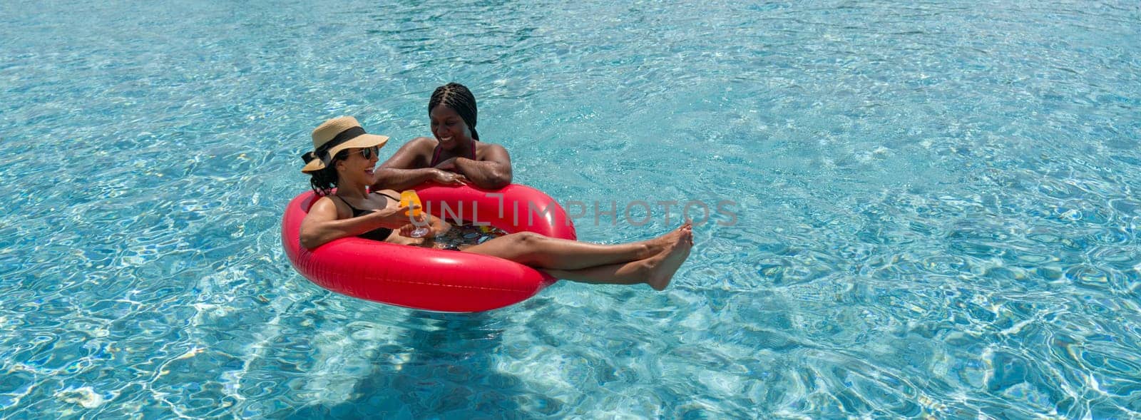 Two ladies in bikini are joyfully floating together on a big inflatable ring in blue water.