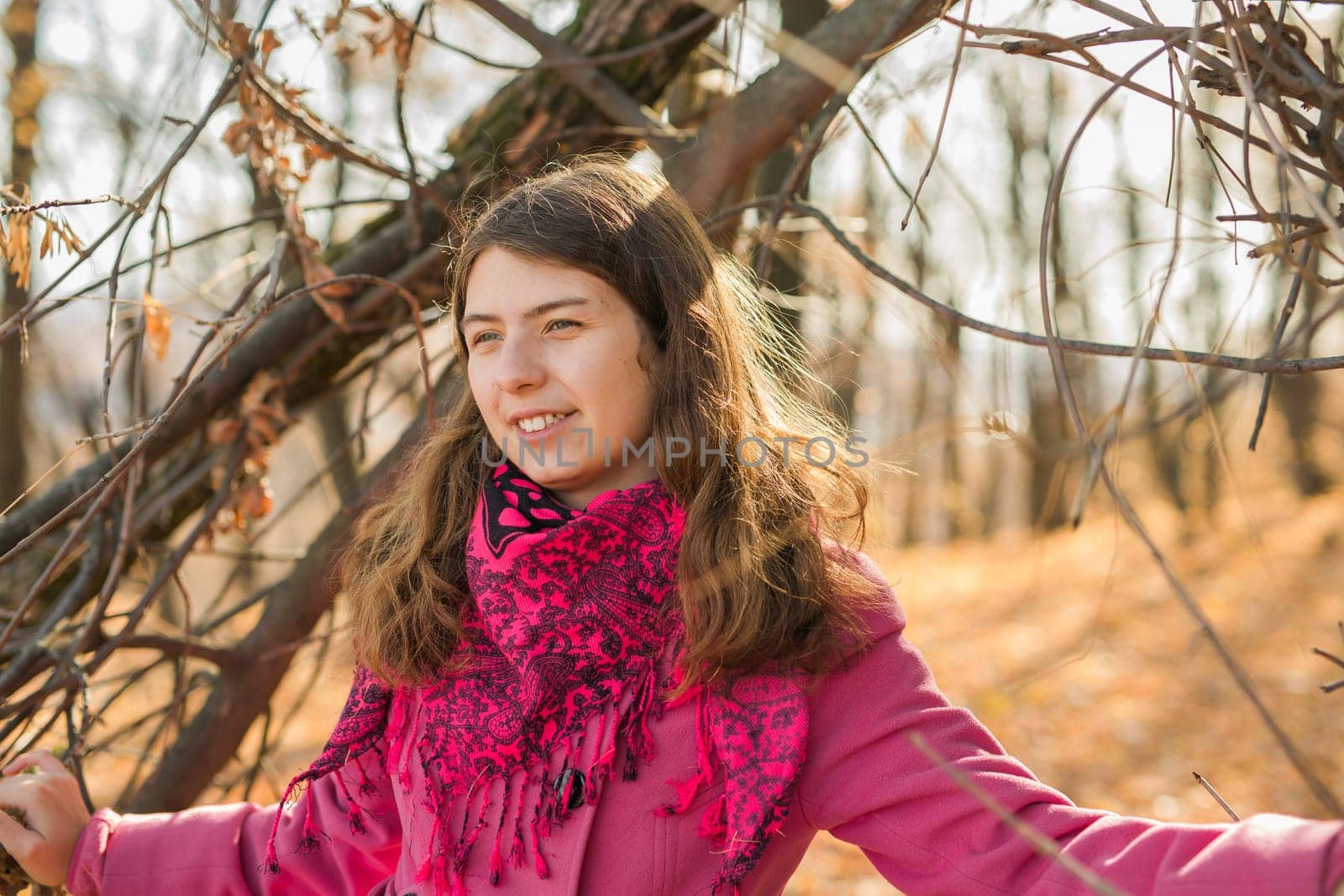 Cheerful young woman with pink scarf relaxing at park with yellow trees in background. Smiling beautiful girl enjoying warm sunny weather in autumn season. Happy pretty natural woman laughing in fall season