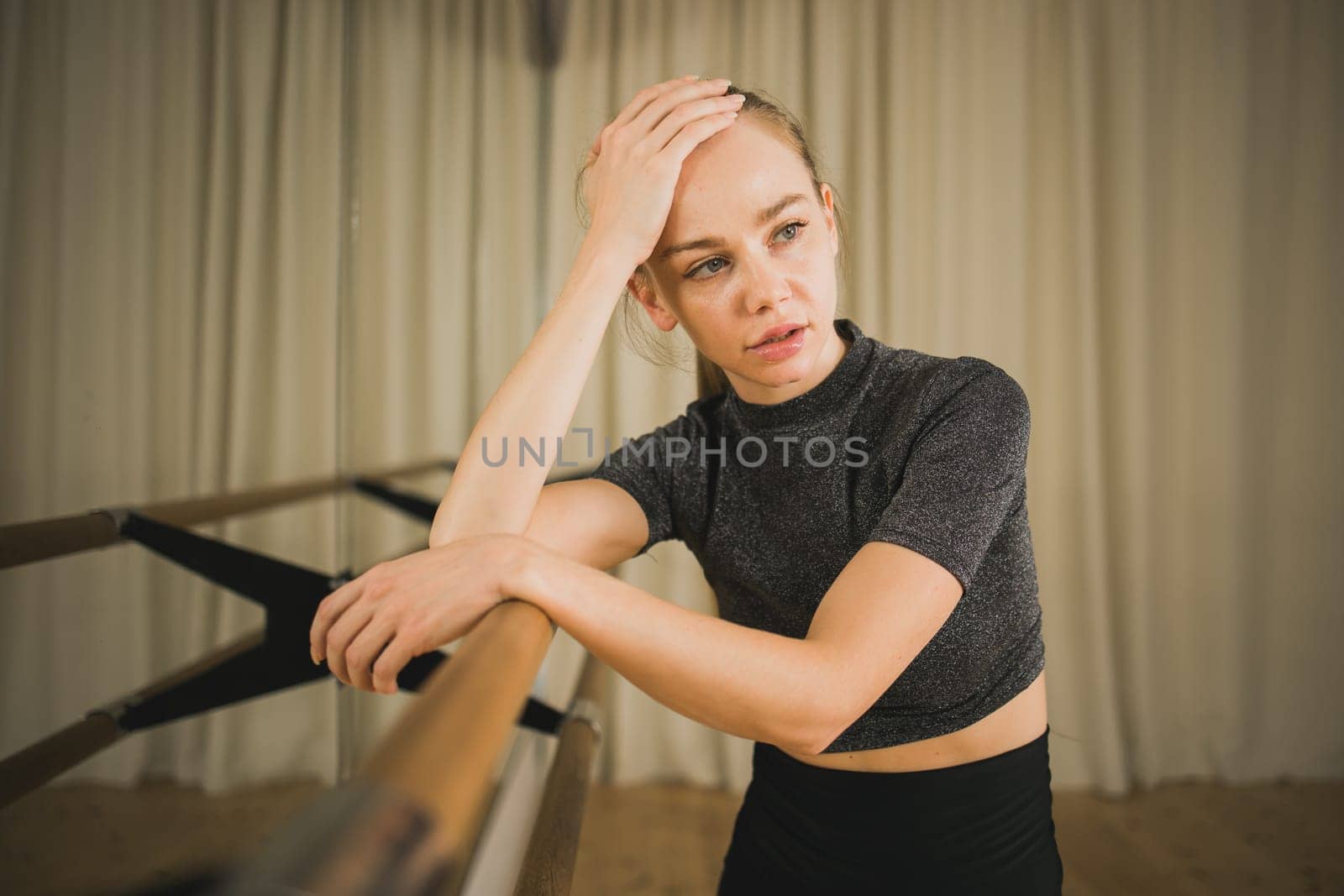 Upset ballerina in training suit stands near ballet barre and looks at the mirror in dance studio. Woman prepares for performance. Ballet dancer doing gymnastic exercises. Classical ballet school.